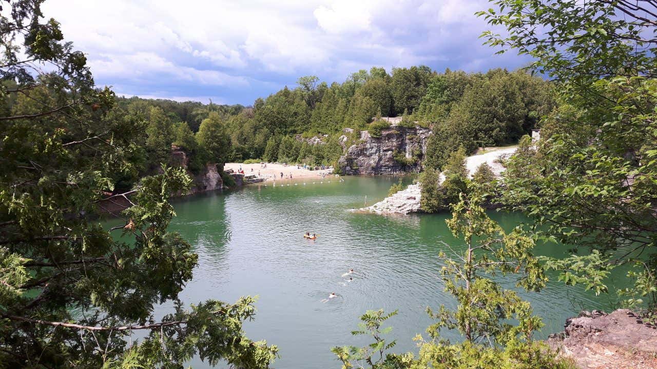 An overhead view of Elora Quarry, a natural swimming hole with green water surrounded by high rock cliffs and dense green trees. People are swimming in the water and relaxing on a sandy beach area.