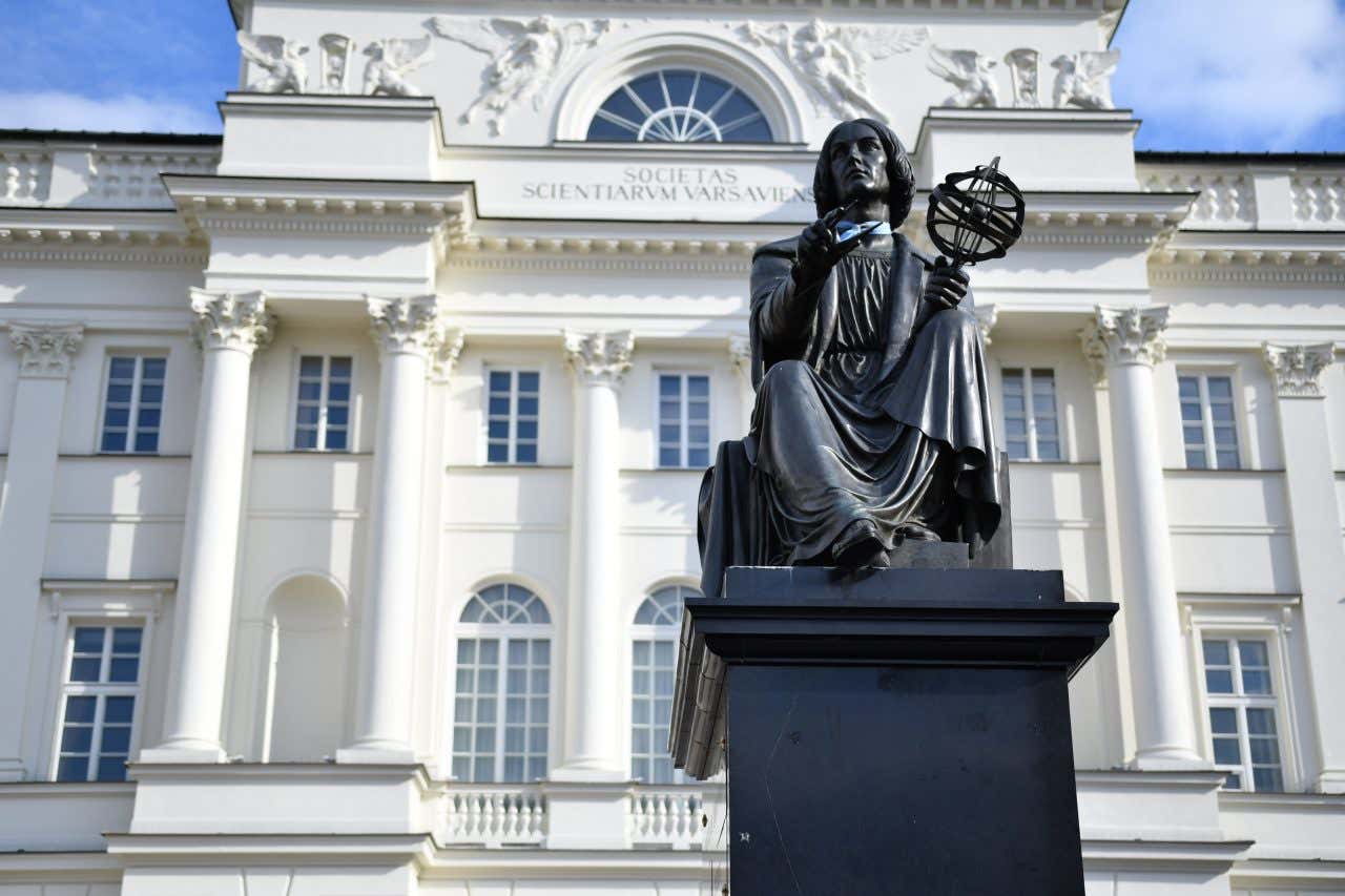 La statua in pietra nera di Copernico vista dal basso. È ritratto seduto su uno scranno mentre tiene in mano un compasso e un astrolabio, alle sue spalle la facciata di un palazzo bianco in stile neoclassico, ovvero quello della Società scientifica polacca