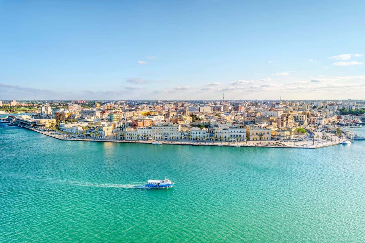 Veduta aerea di Brindisi ripresa dal mare in una giornata di sole, con il porto visibile sul lato destro dell’immagine e diverse navi attraccate alle banchine. Al centro e a sinistra si estende il centro storico con edifici chiari. In primo piano, un motoscafo sfreccia sull’acqua lasciando una scia bianca