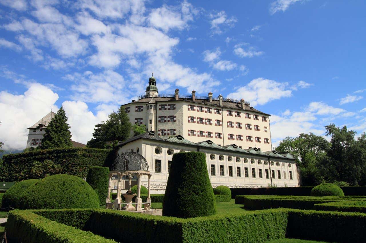 Il Castello di Ambras a Innsbruck, Austria, edificio rinascimentale bianco con tetto scuro e numerose finestre, circondato da giardini formali con siepi potate e un elegante padiglione in primo piano, sotto un cielo azzurro e parzialmente nuvoloso