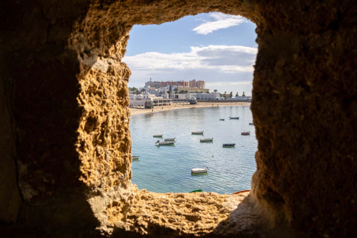 Una ventana de piedra perteneciente a un castillo con vistas al mar, una playa y varios barcos de pescadores