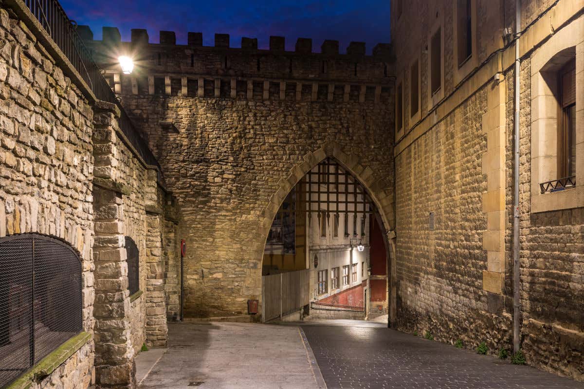 Una calle de aspecto medieval iluminada por la noche y con un gran pórtico con una verja metálica
