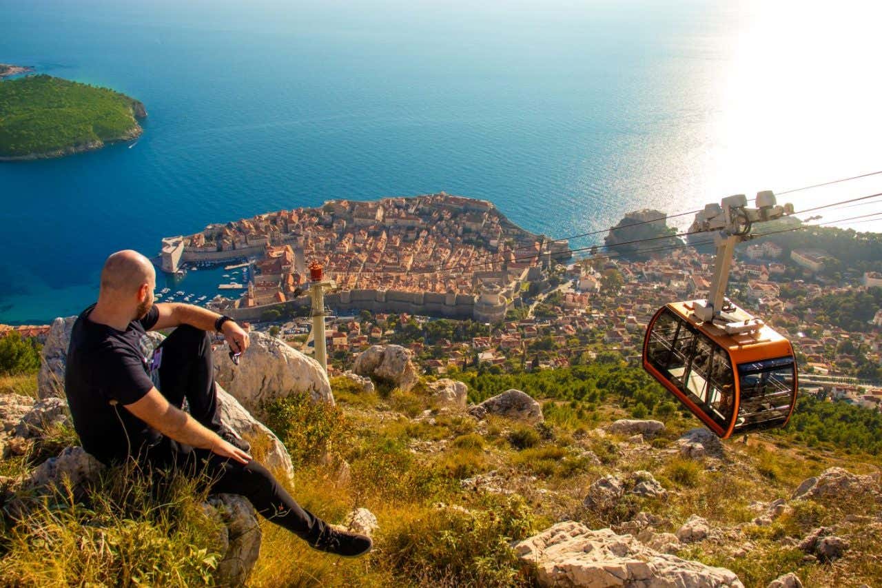 Un hombre sentado en un mirador de un monte rocoso ve hacia una ciudad y un teleférico