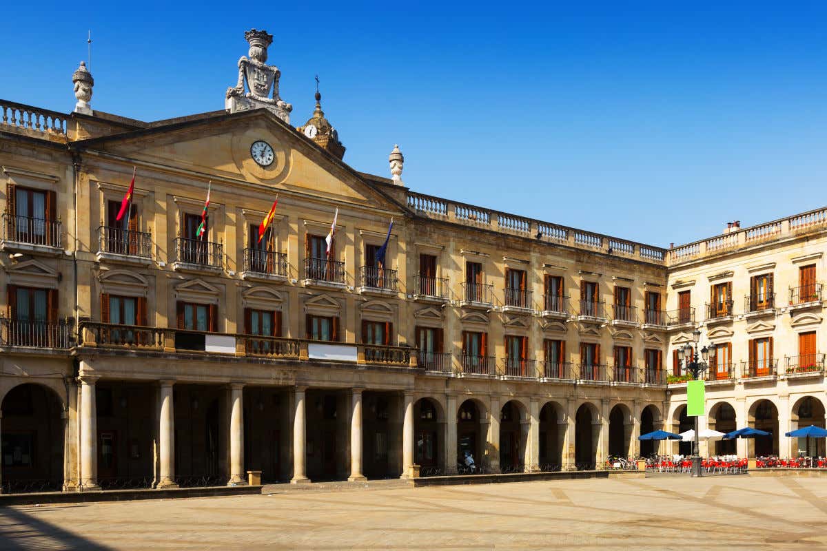 Ayuntamiento de Vitoria con varias banderas y un reloj en su fachada, estando el edificio en una plaza cuadrada con múltiples soportales