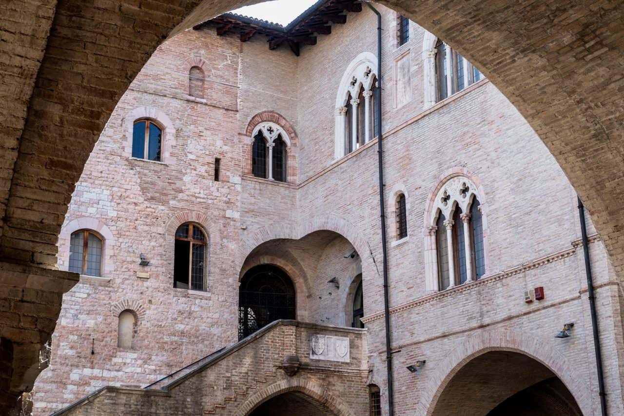 Veduta del cortile interno di Palazzo Trinci a Foligno, con archi ogivali in primo piano, scala monumentale in mattoni che conduce a un portale ad arco, e finestre gotiche bifore e trifore decorate con colonnine e archi trilobati sulle pareti in pietra chiara