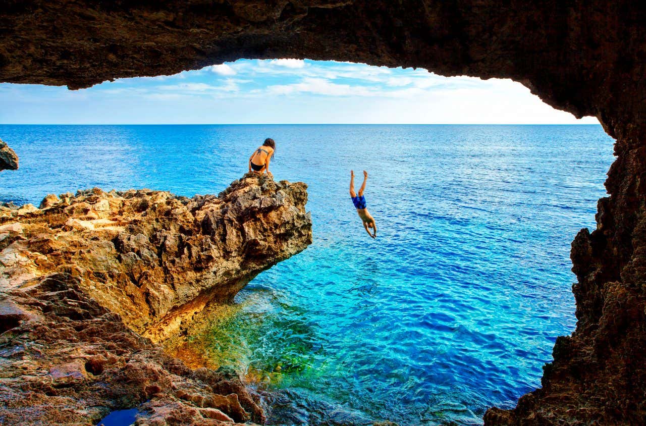 Due giovani su una scogliera rocciosa a picco sul mare limpido vicino a Protaras, Cipro: uno si tuffa in acque turchesi da una sporgenza, mentre l’altra osserva seduta, incorniciati dall’ombra di una grotta naturale in primo piano