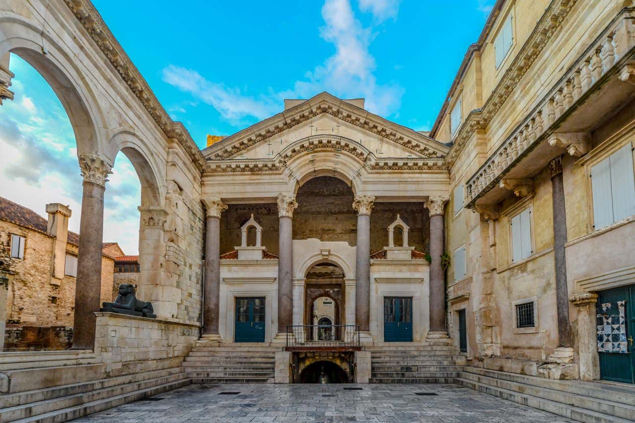The Peristyle in Diocletian's Palace, Split, Croatia, an ancient Roman courtyard with large stone columns and arches. The central building features a grand entrance with a balcony and two large blue doors.