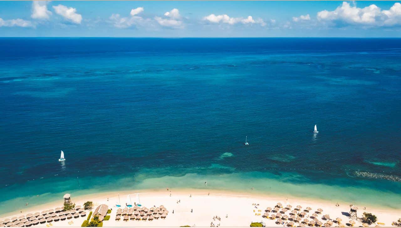 An aerial view of Montego Bay with sun beds seen on the beach, and some boats in the deep blue ocean.