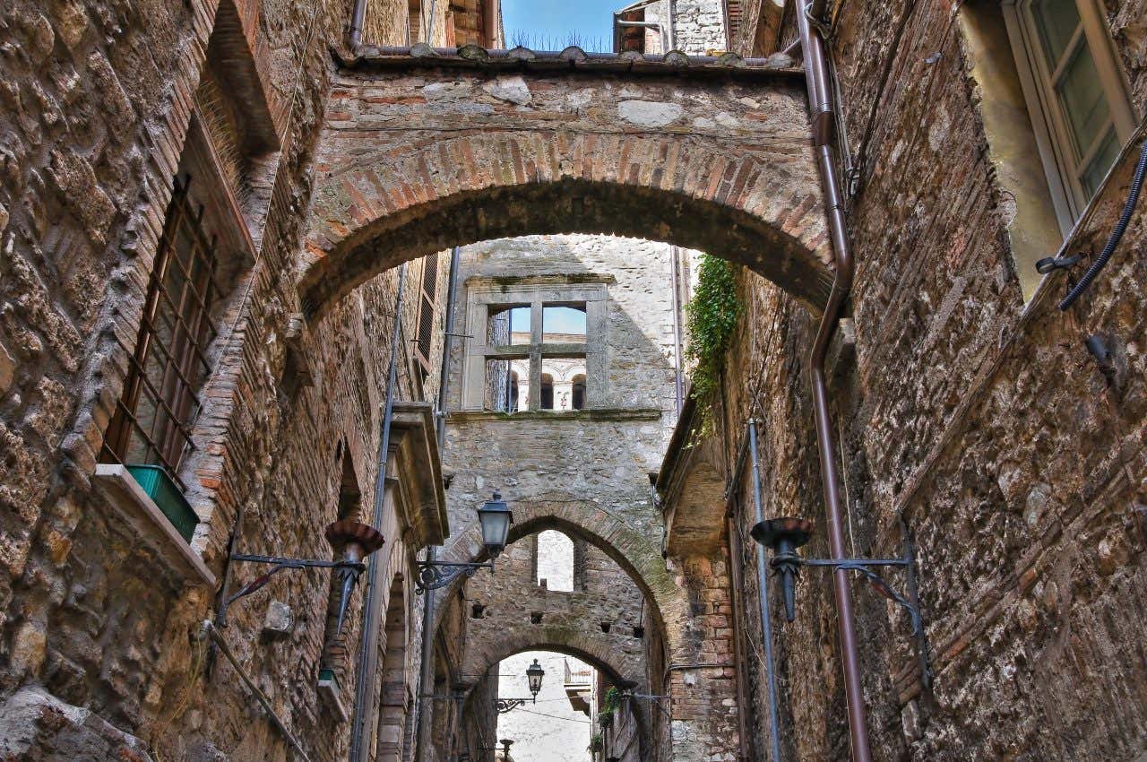Vicolo medievale in pietra a Narni, Umbria, con archi in mattoni, passaggi coperti tra edifici, finestre antiche e lampioni in ferro battuto, visto dal basso verso l’alto in una giornata soleggiata