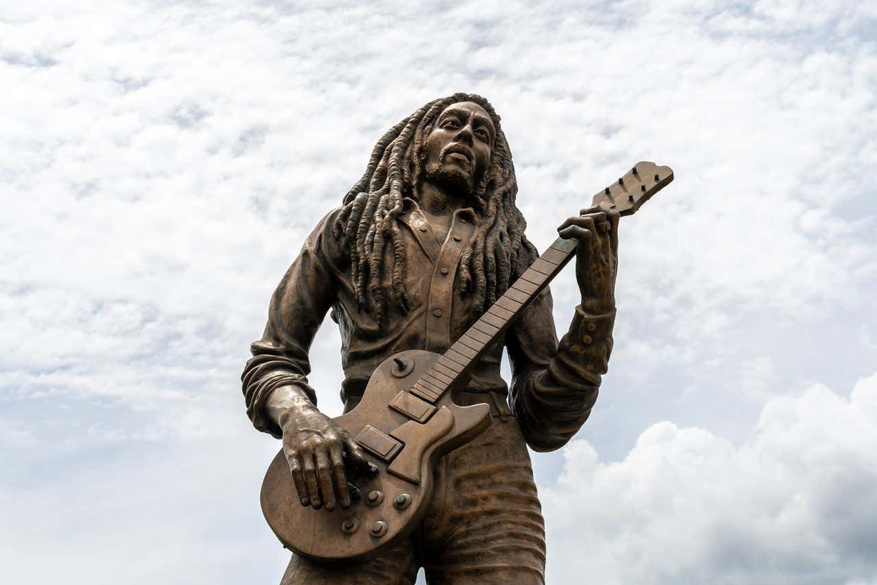 A bronze statue of Bob Marley, depicted holding a guitar with his dreadlocks. The statue is seen from a low angle against a cloudy sky.