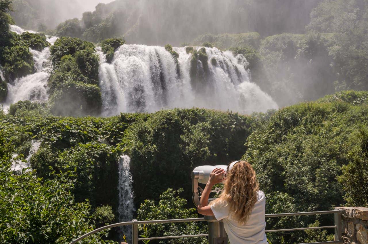 Una ragazza osserva la possente cascata delle Marmore, che scorre in mezzo alla boscaglia, da un monocolo fisso installato su un belvedere in una giornata estiva di sole