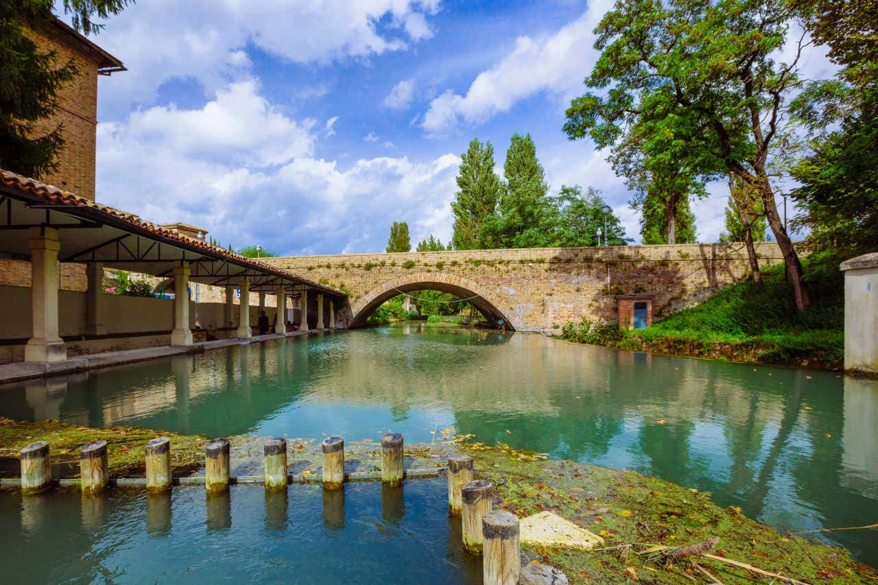 Il lavatoio dell’Accolta a Bevagna in una giornata di sole, con vasca in pietra riempita da acqua limpida e riflessi dorati, circondata da muretti rustici e vegetazione; sullo sfondo, case in pietra con persiane chiuse e un ponte ad arco in mattoni che attraversa il canale