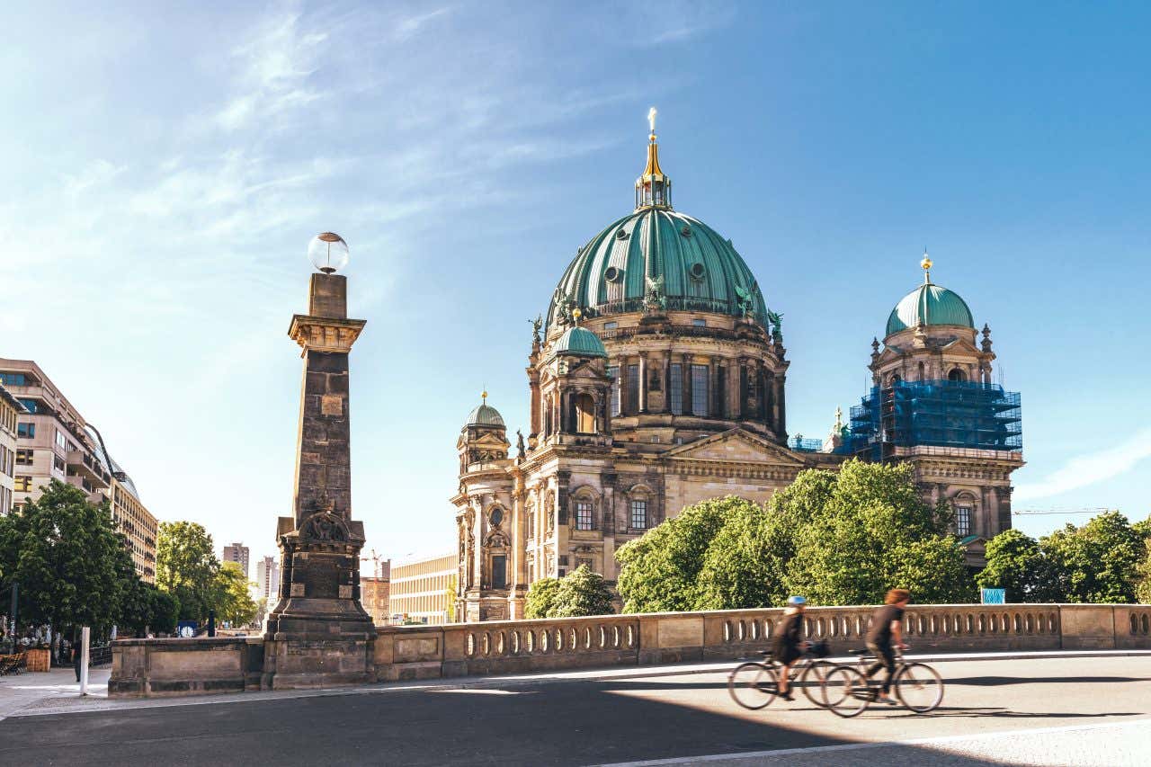 Two people blurred in motion cross a bridge in front of the Berlin Cathedral on a sunny day in Berlin, one of the best cities for biking