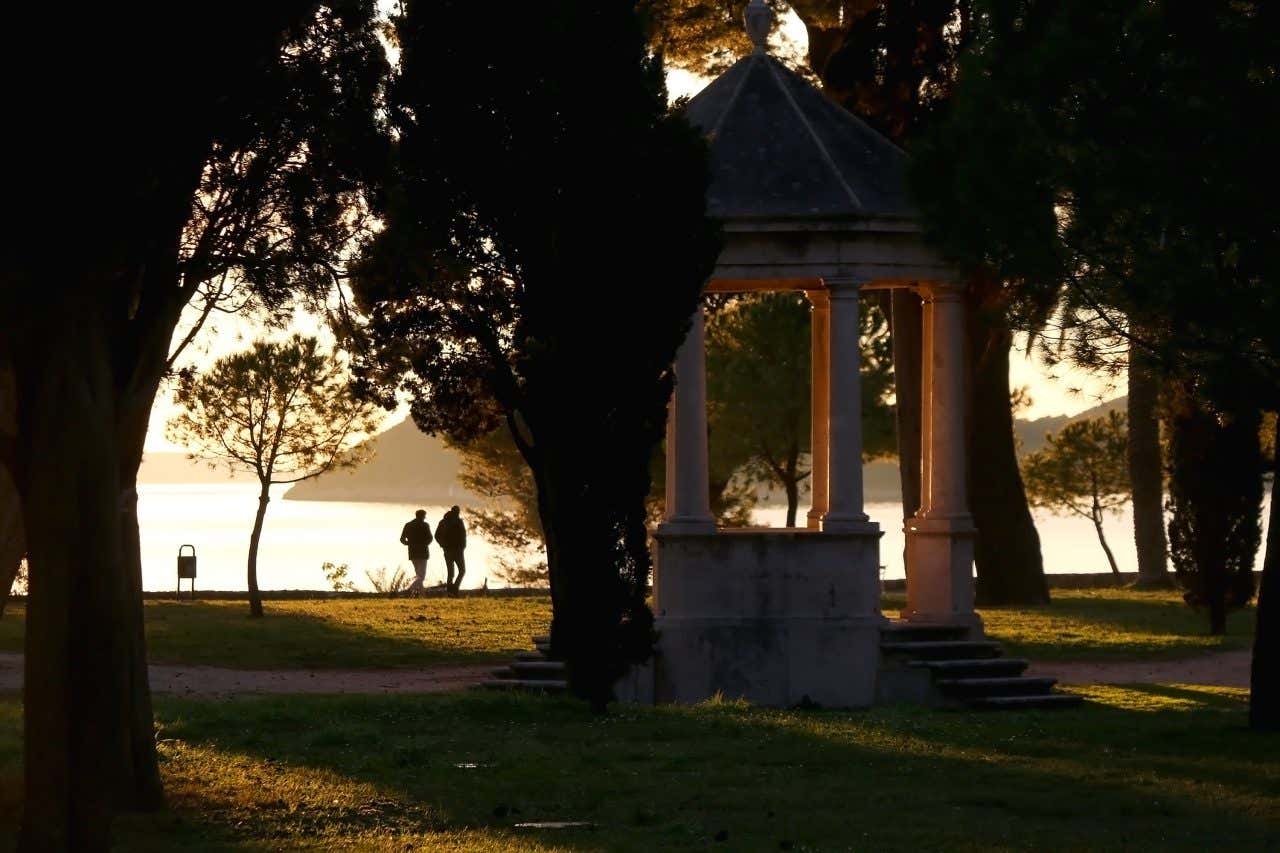 A gazebo in Sustipan Park, Split, Croatia, framed by dark trees. Two people are walking in the background along the waterfront, with the sea visible under a bright sky, likely during sunrise or sunset.