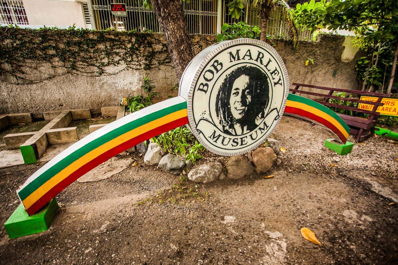 The entrance sign for the Bob Marley Museum, featuring a large circular emblem with Bob Marley's portrait and "BOB MARLEY MUSEUM" written around it. The emblem is supported by two arching structures painted in red, yellow, and green.