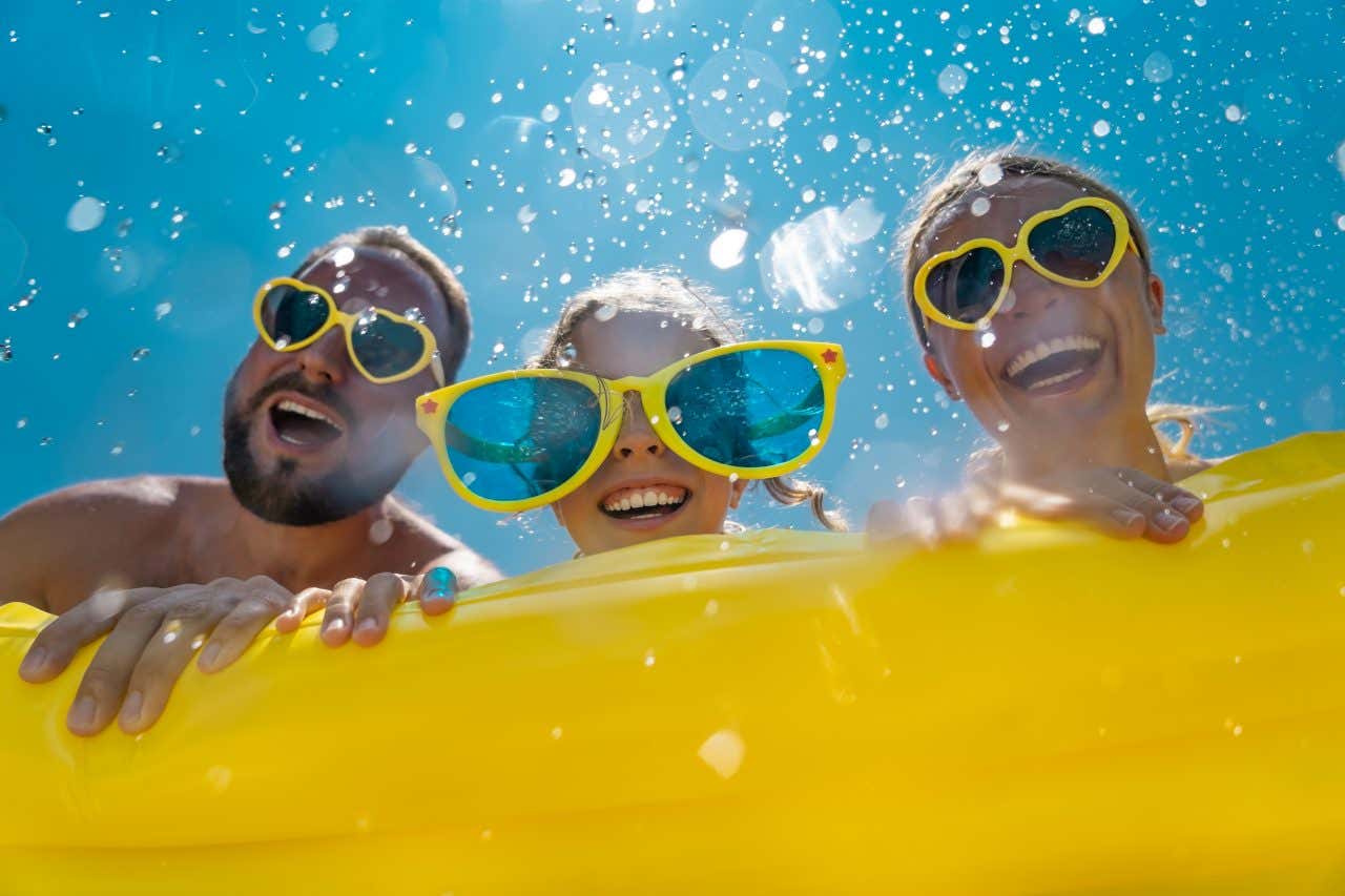 Foto dal basso di una famiglia composta da un uomo, una donna e una bambina. Sorridono mentre indossano occhiali da sole colorati. Si divertono in un parco acquatico, appoggiati a un grande gonfiabile giallo con schizzi d'acqua che volano intorno sotto un cielo blu