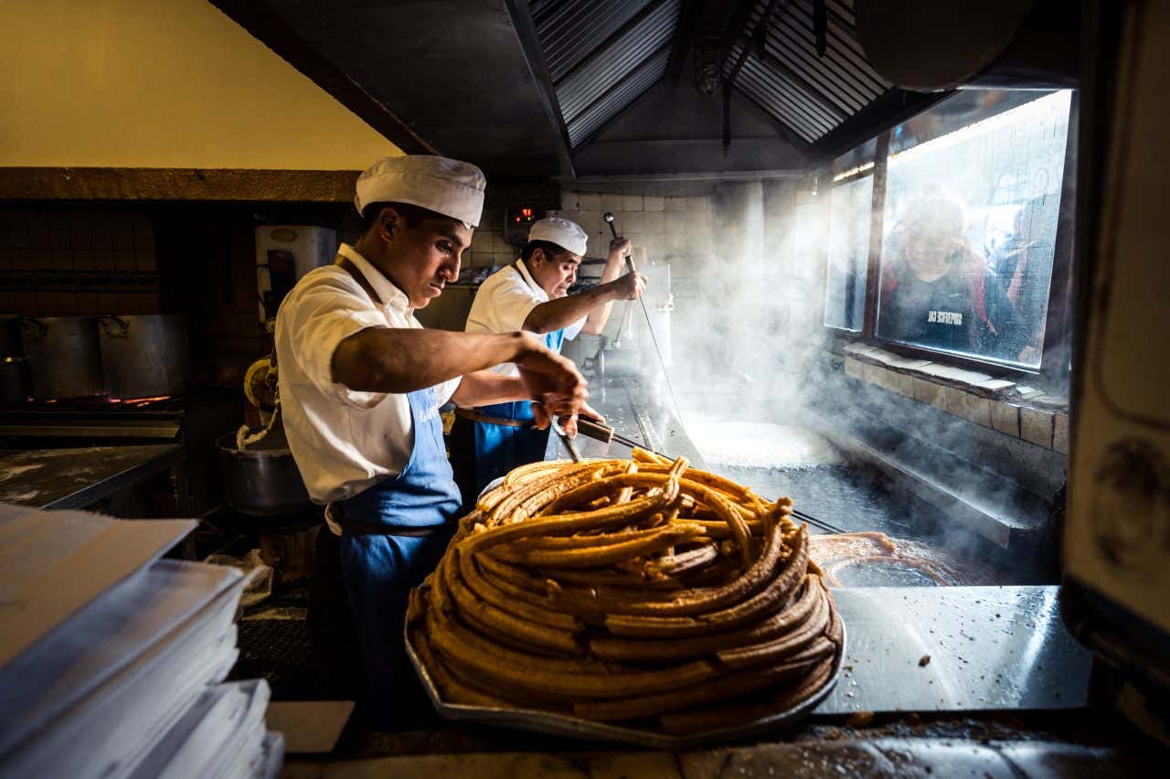 Two chefs are working in a kitchen, preparing churros. One chef is holding a large pile of golden-brown churros on a tray in the foreground, while another works with cooking equipment in the smoky background.
