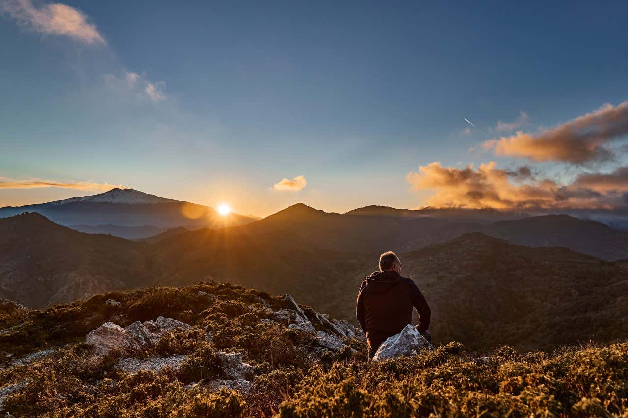 Un uomo di spalle osserva un panorama montuoso dalla cima dell'Etna durante il tramonto, che tinge il paesaggio di sfumature arancioni