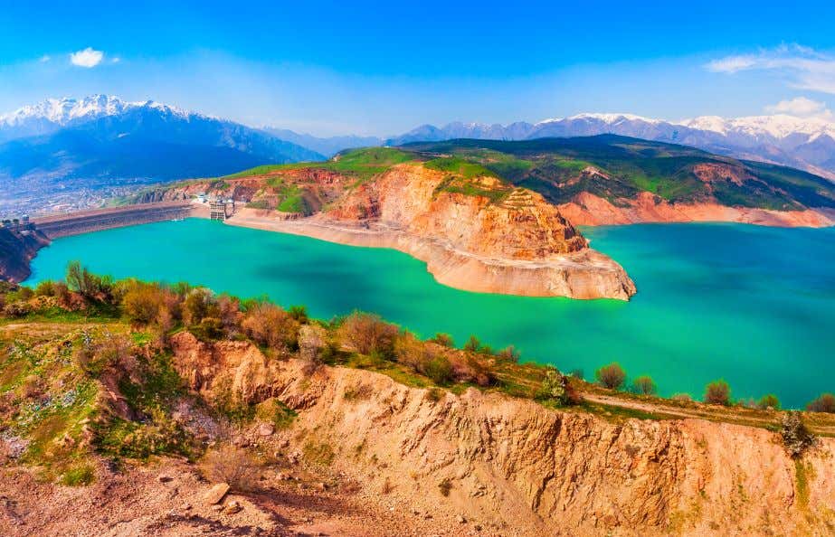 A view of Lake Charvak in Uzbekistan, seen in Spring under a clear blue sky, with snow-capped mountains in the distance.