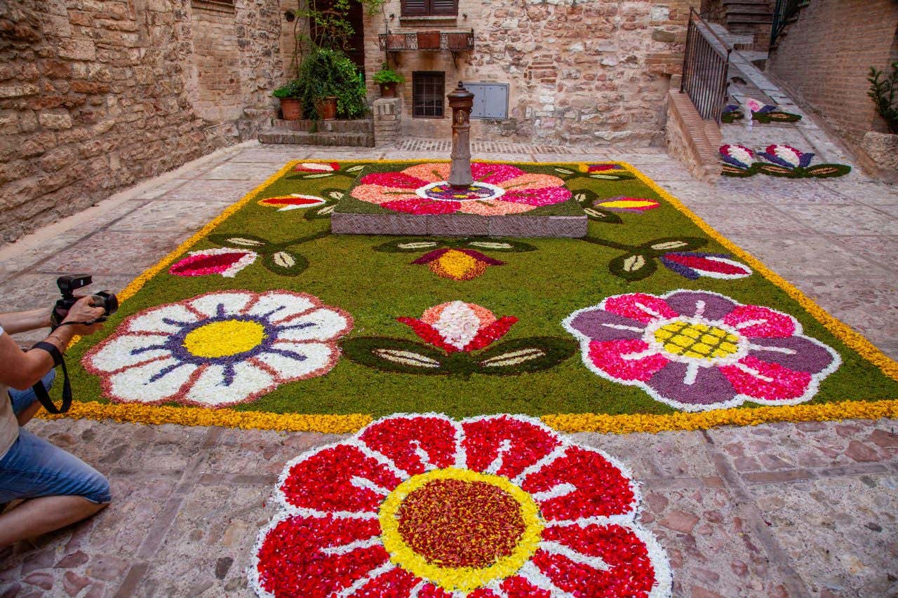 Decorazione floreale artistica a tappeto in una piazzetta di Assisi, con motivi di grandi fiori colorati realizzati con petali su sfondo verde, disposti attorno a una colonna in pietra al centro; sullo sfondo, edifici in pietra con vasi di piante e gradini laterali, mentre un fotografo scatta un'immagine in primo piano