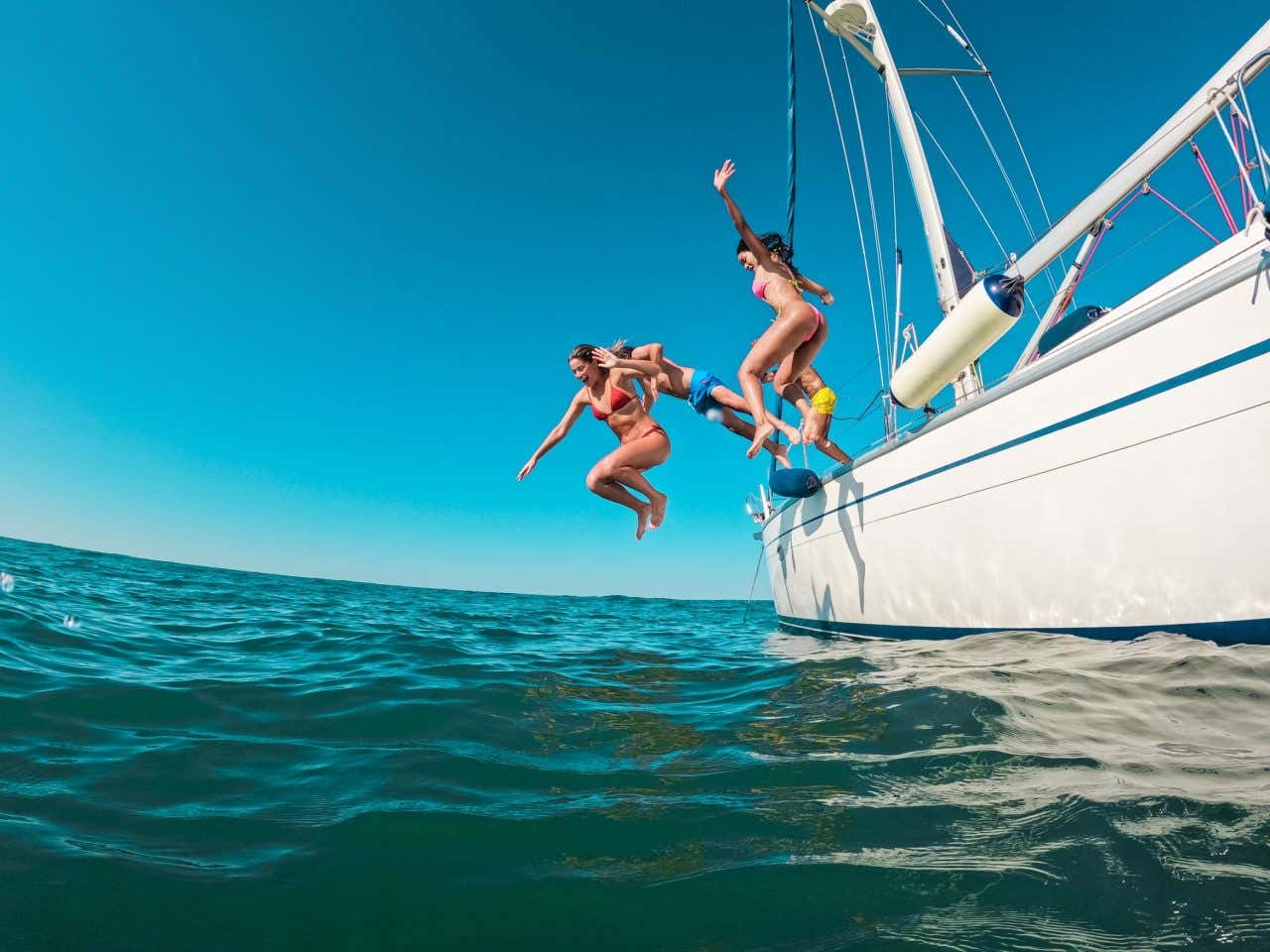 Due ragazze e due ragazzi in costume da bagno nel momento in cui si stanno tuffando nel mare da una barca bianca in una giornata estiva senza nuvole