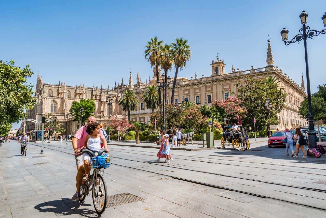 A father and daughter ride a bike in front of the classical building in Seville, the most bike friendly city in Spain, on a clear sunny day