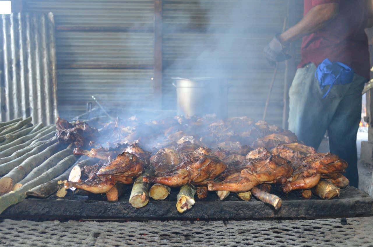 Whole chickens are cooking on a large outdoor grill, with smoke rising from the meat. The grill is made of metal and wood, and a person is partially visible in the background.