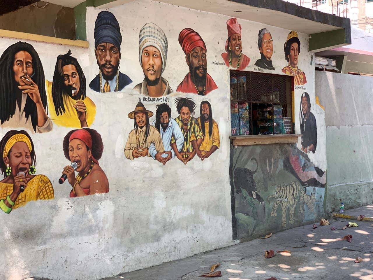 A wall mural depicting multiple portraits of famous Jamaican musicians and cultural figures, including Bob Marley, against a light-colored background. Some figures are shown playing instruments or singing.