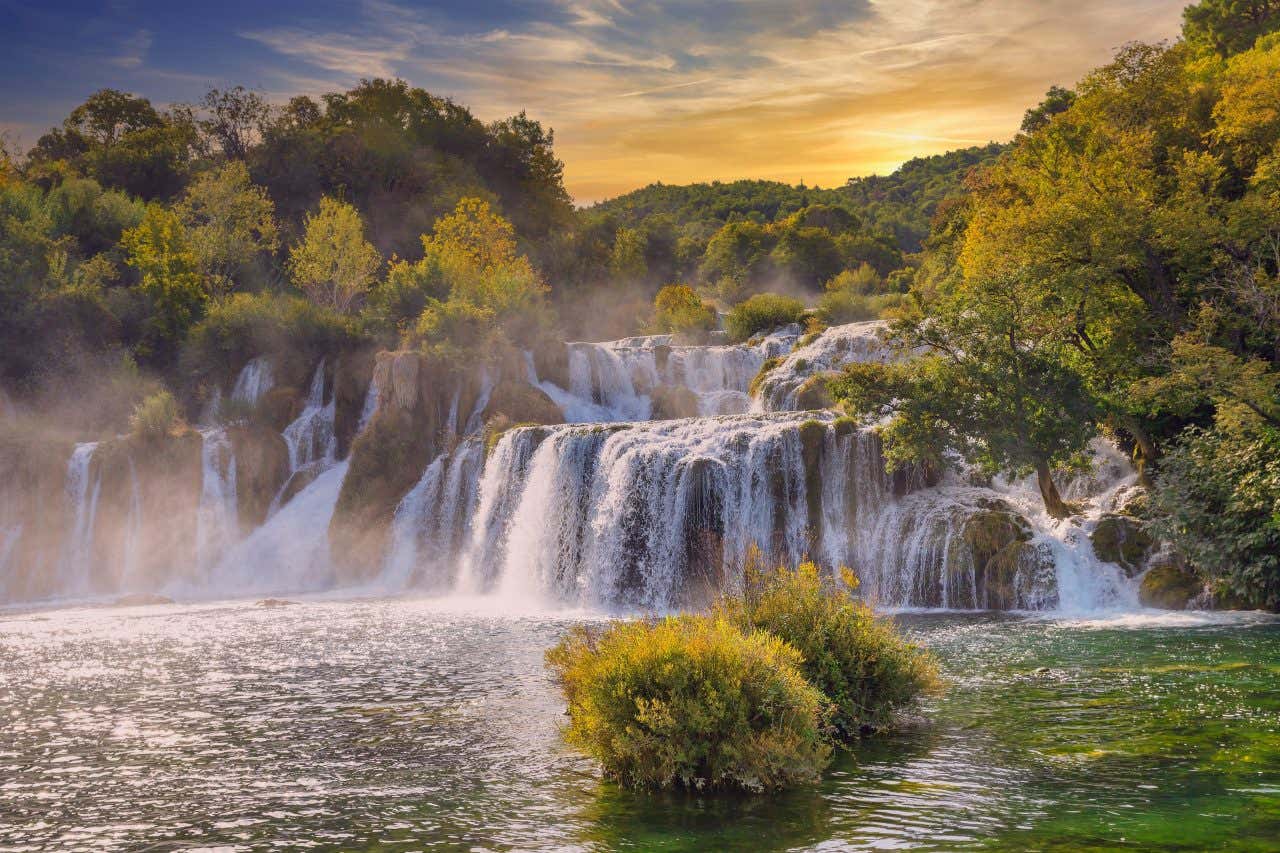 Skradinski Buk, a wide, multi-tiered waterfall in Krka National Park, Croatia. Water cascades over limestone formations, surrounded by lush green and autumn-colored trees under a sky with hints of orange and blue.