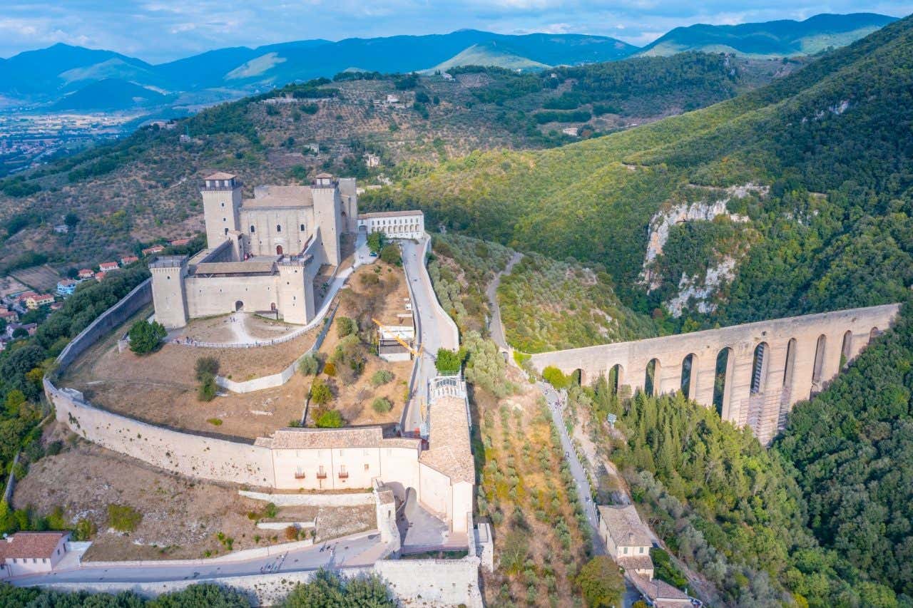 Veduta aerea soleggiata della Rocca Albornoziana, imponente fortezza medievale in cima al colle Sant’Elia, con le sue sei torri e le mura spesse; sullo sfondo scorre il monumentale Ponte delle Torri ad arcate che collega la rocca al bosco di Monteluco, con i tetti del centro storico e la verdeggiante vallata spoletina sottostante