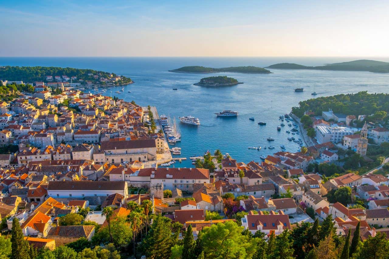 An elevated view of Hvar Town on Hvar Island, Croatia, showing a densely built town with red rooftops extending to a large harbor filled with boats. Several islands are visible in the blue Adriatic Sea in the distance under a sunny sky.