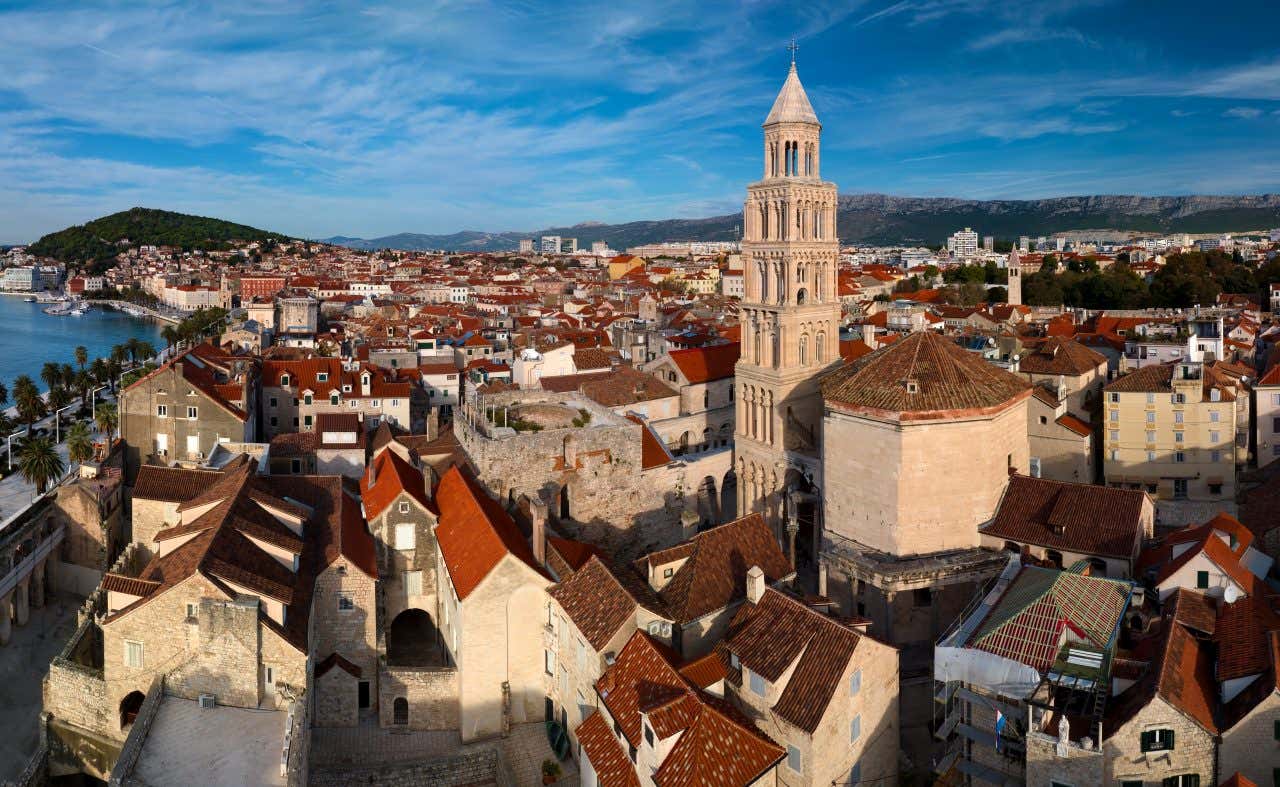 An aerial view of Split, Croatia, showing a dense cluster of red-roofed buildings and the prominent bell tower of Split Cathedral. The Adriatic Sea and mountains are visible in the background under a blue sky.