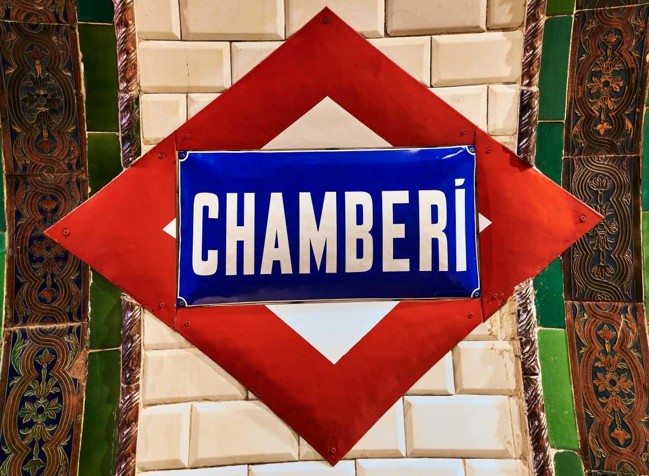 Insegna in stile retrò della stazione della metropolitana di Chamberí a Madrid, Spagna. Il cartello è blu con lettere bianche e incorniciato da una forma romboidale rossa su sfondo di piastrelle bianche e dettagli decorativi in ceramica verde e marrone ai lati