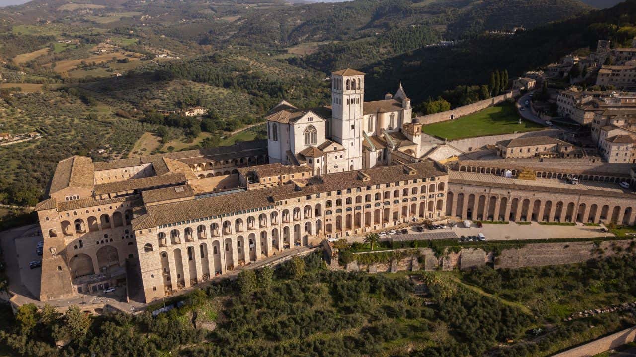 Foto aerea della Basilica di San Francesco d’Assisi: visibili la basilica superiore in stile gotico con rosone centrale e campanile romanico, e quella inferiore romanica con contrafforti massicci; il complesso si erge su un promontorio con arcate del Sacro Convento a sostegno