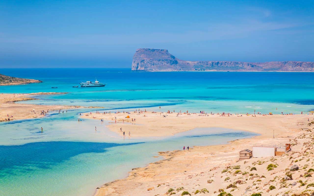 Vista aerea mozzafiato della laguna di Balos, Creta: la sabbia bianca e rosa si estende tra acque turchesi poco profonde e mare blu intenso, mentre un promontorio roccioso emerge sullo sfondo sotto un cielo sereno. Sulla spiaggia in primo piano si vedono alcuni bagnanti