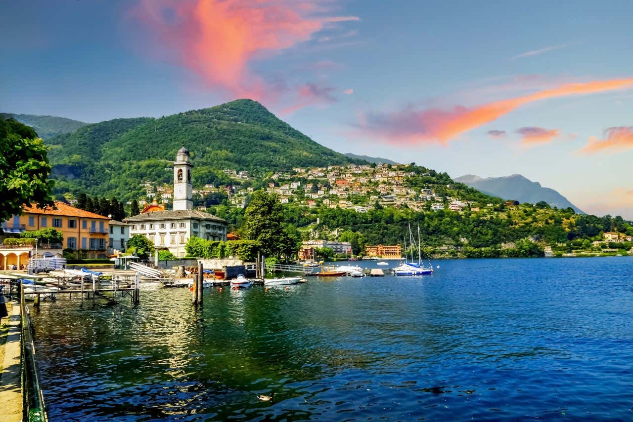 A town on the shore of Lake Como, Italy, with buildings and a prominent white bell tower. Boats are docked along the waterfront, and a forested mountain rises behind the town under a sky with pink clouds.