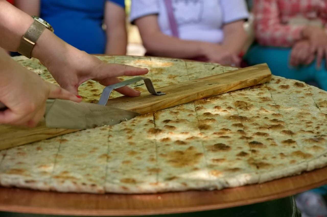 A soparnik, a traditional Croatian pie being cut with a knife and a wooden plank, with people watching in the background.