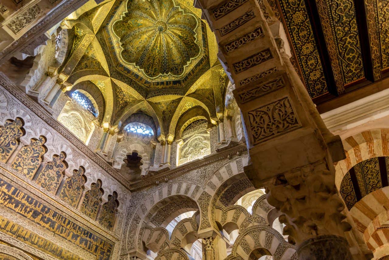 Vista dal basso verso l’alto all’interno della Moschea-Cattedrale di Córdoba: le arcate a ferro di cavallo con alternanza di mattoni rossi e pietra chiara si sovrappongono in sequenza, sostenute da colonne scolpite, mentre il soffitto decorato rivela cupole e volte in stile moresco