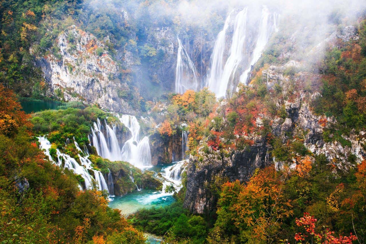 A panoramic view of Plitvice Lakes National Park in Croatia, showcasing multiple waterfalls cascading between terraced, turquoise lakes. The surrounding landscape is covered in dense foliage, displaying vibrant autumn colors and some mist.