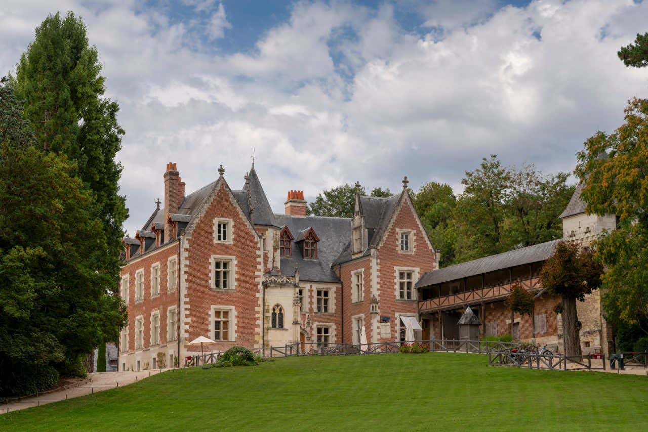 Vista del Château du Clos Lucé immerso nei toni autunnali direttamente dal giardino: la facciata in mattoni rossi e pietra chiara emerge tra le fronde degli alberi autunnali, mentre il cielo è coperto da nuvole gonfie