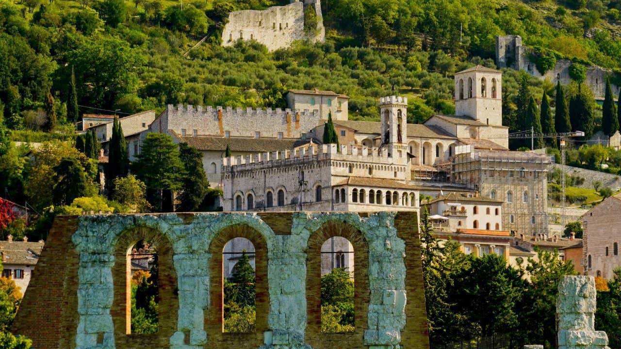 I palazzi rinascimentali e medievali del centro storico di Gubbio abbarbicati alle pendici di una verde collina. In primo piano i resti di un anfiteatro romani a valle.