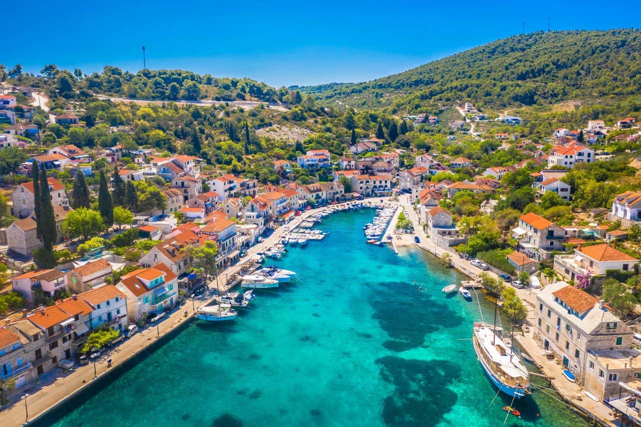 A shot of the port of Brač, Croatia with turquoise waters, red-roofed buildings and a green mountain view in the background.