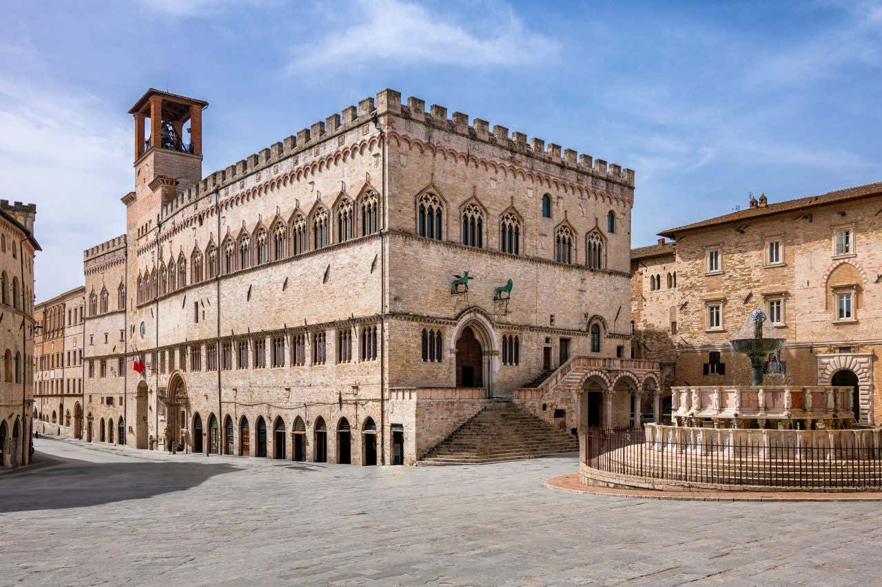 Piazza IV Novembre a Perugia vista inquadrando la Fontana Maggiore centrale con le sue due vasche marmoree e la tazza bronzea saturnina; sullo sfondo, la facciata gotica del Palazzo dei Priori in pietra, con trifore al piano superiore, portale trilobato fiancheggiato da coppe bronzee del grifo e leone, scalinata a ventaglio che conduce alla Sala dei Notari