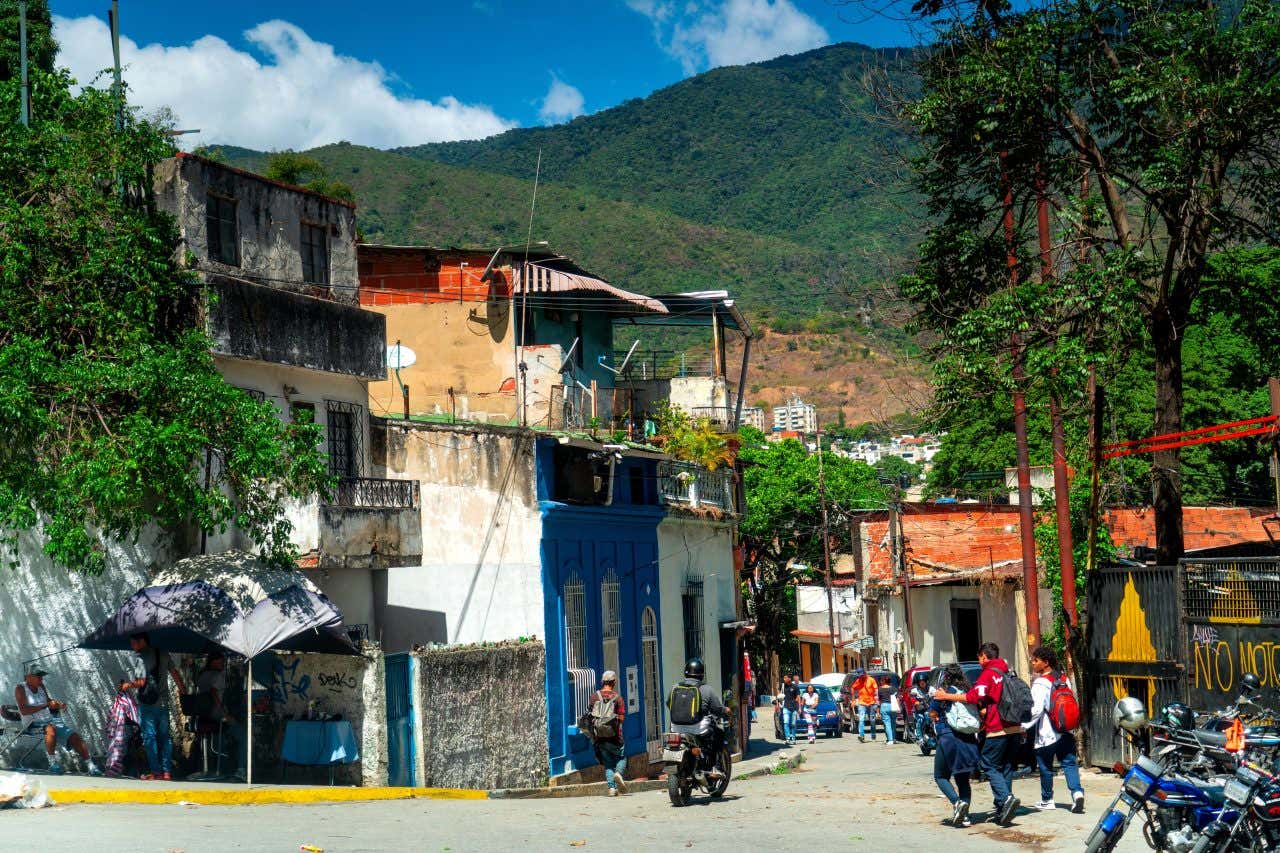 A street in a residential neighborhood in Caracas, Venezuela, with buildings of various colors and conditions lining the road. People are walking and a motorcycle is present, with a large green mountain rising in the background under a blue sky.