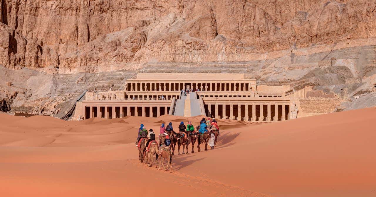 Carovana di turisti su cammelli che attraversa il deserto arancione verso il Tempio di Hatshepsut, situato ai piedi di una scogliera rocciosa nei pressi di Luxor, in Egitto