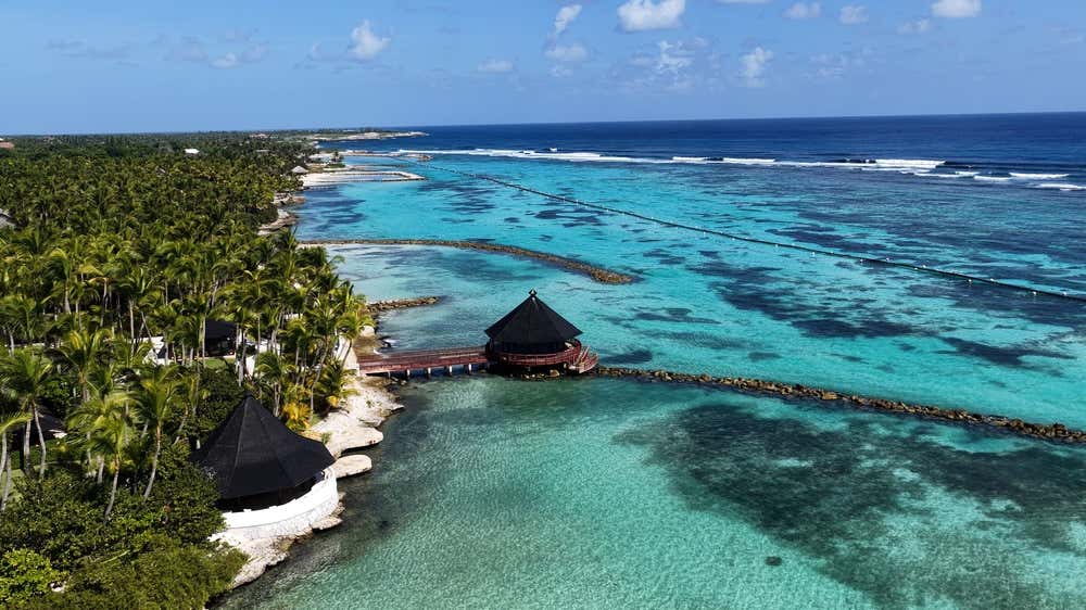 Foto panoramica dall'alto della spiaggia Playa Blanca a Punta Cana, con sabbia bianca, mare turchese e un bosco di palme e moli di pietra che entrano in acqua