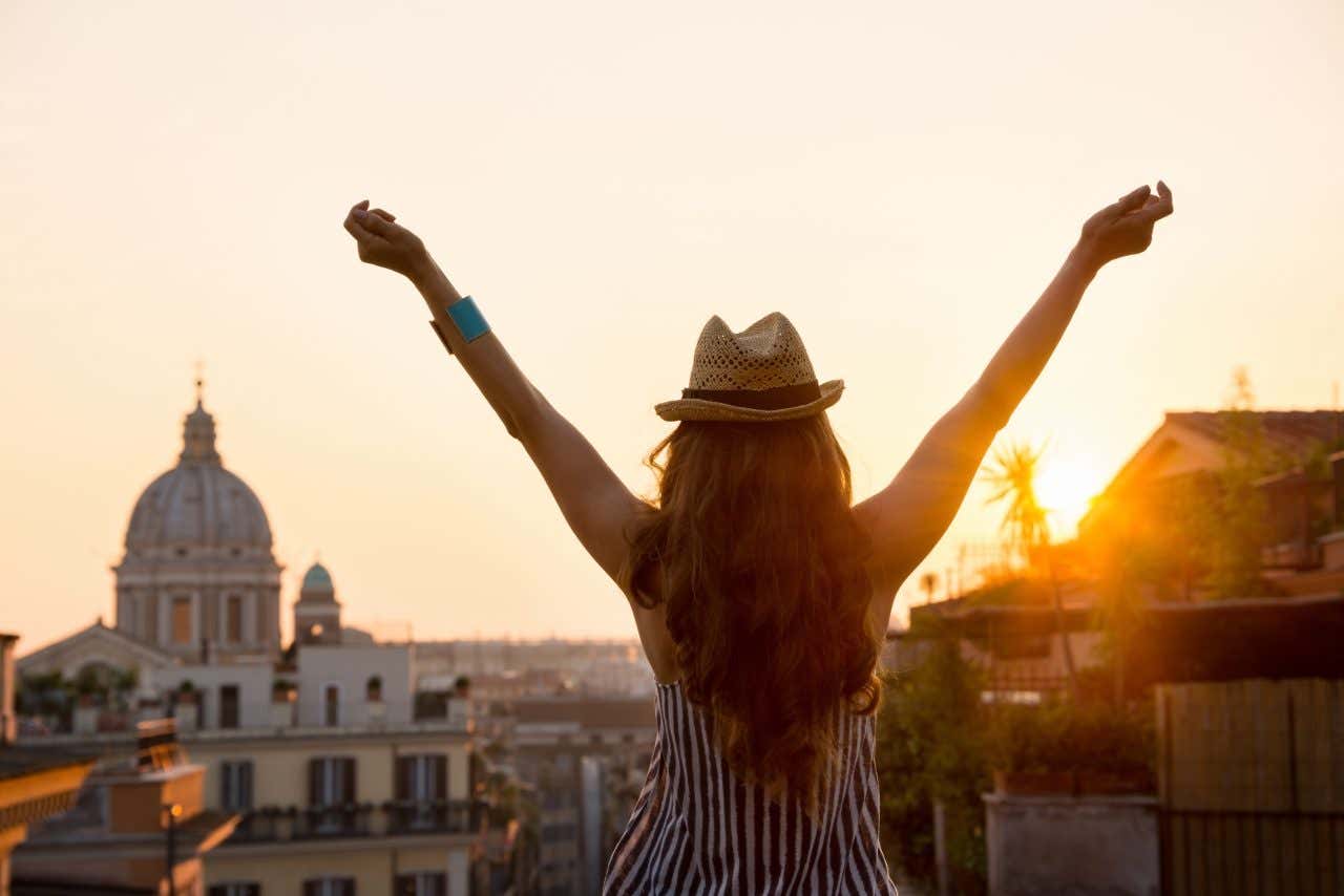 Una ragazza dai capelli lunghi e castani di spalle e con un cappello di paglia, alza le braccia al cielo mentre ammira il tramonto sul centro storico di Roma. Sul fondo si intravede una cupola di una chiesa barocca