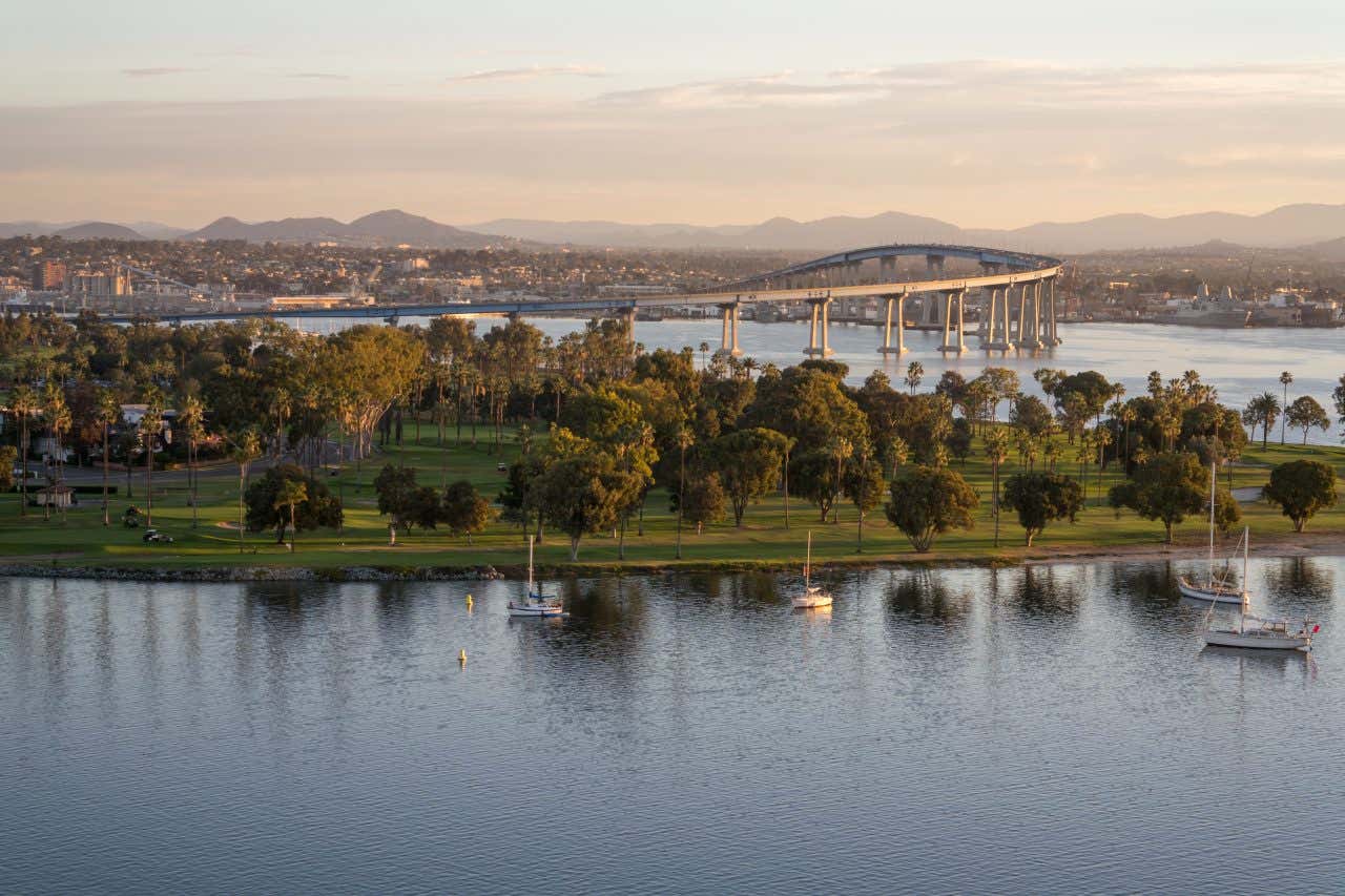 Veduta panoramica di San Diego al tramonto, con il Coronado Bridge che si estende sopra la baia, circondato da un parco verde con palme, alberi e barche a vela ormeggiate sull’acqua tranquilla in primo piano. Sullo sfondo, si intravede la città e le colline sotto un cielo dorato