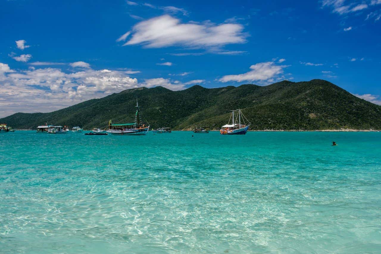 Água turquesa repleta de barcos com montes verdes ao fundo em um dia ensolarado