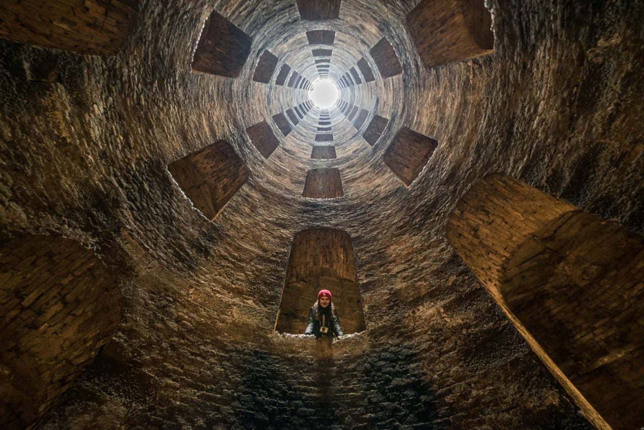 Persona con cappello rosso che si affaccia da una finestra interna all'interno del Pozzo di San Patrizio a Orvieto, visto dal basso verso l'alto con luce naturale che entra dalla sommità del pozzo cilindrico in pietra, circondato da una scala elicoidale e archi simmetrici